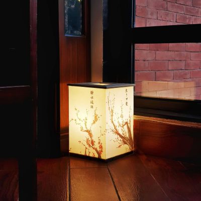 A square, Japanese-style Hako lamp with cherry blossom patterns and kanji characters, glowing warmly on a wooden floor in a dimly lit corner next to a window with a brick wall outside.