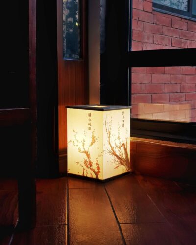 A square, Japanese-style Hako lamp with cherry blossom patterns and kanji characters, glowing warmly on a wooden floor in a dimly lit corner next to a window with a brick wall outside.