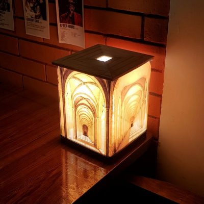 A square Demi lamp glowing warmly with an illusion of a vaulted hallway on its sides, sitting on a reflective wooden surface against a brick wall with posters.