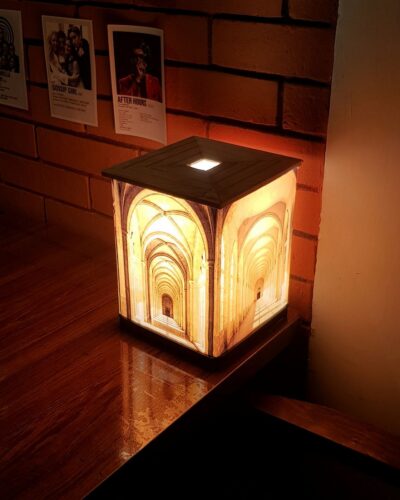 A square Demi lamp glowing warmly with an illusion of a vaulted hallway on its sides, sitting on a reflective wooden surface against a brick wall with posters.